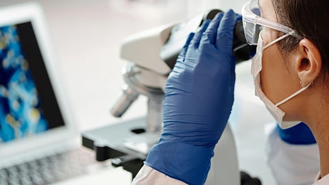 A woman in a lab coat examines a specimen through a microscope in a laboratory setting.