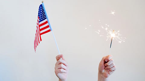 Two hands, one holding a small American flag and the other holding a lit sparkler.
