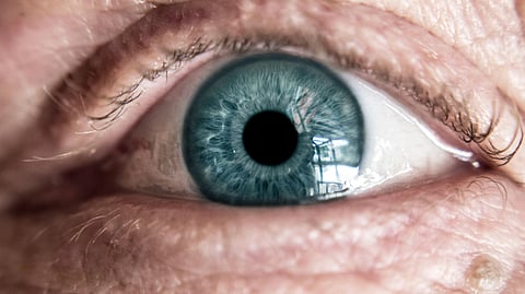 A close-up photograph of a light blue human eye surrounded by detailed skin texture and fine wrinkles.