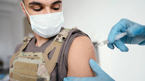 A masked man in a tactical vest receives an injection in his upper arm.
