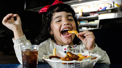 A young girl smiles while eating fries with cheese sauce and drinking a soft drink.
