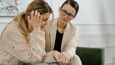 A distressed woman sits with her head in her hand while a counselor gently holds her hand in support.