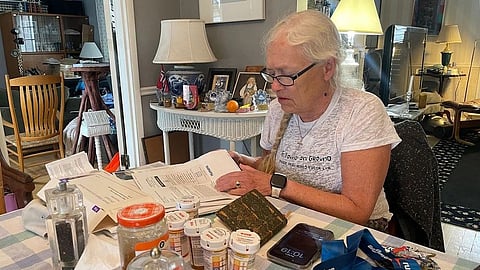An elderly woman reads paperwork at a table with medicine bottles and daily items around her.