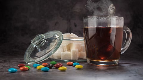 A glass mug of tea sits next to an open jar of sugar cubes, with colorful candies scattered on the table.