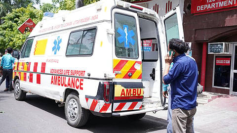 An Advanced Life Support ambulance is parked outside with its rear door open, while a man talks on a phone nearby.
