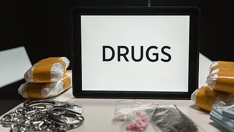 A phone showing “DRUGS” on the screen is surrounded by taped packages, handcuffs, and packets of pills and powder.