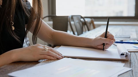 A person writing on paper on a clipboard on a wooden table.