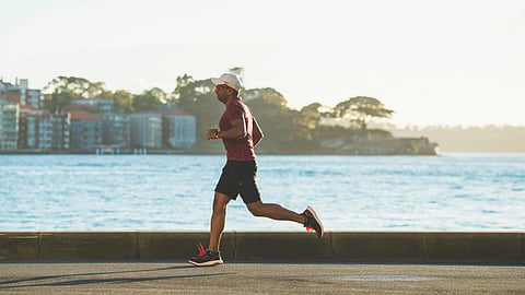 A man runs along a sunny waterfront path near calm water and distant buildings.
