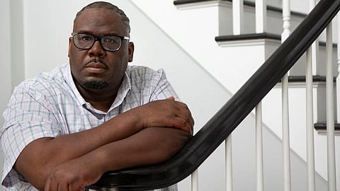 A Black man with glasses and a patterned shirt leans on a staircase banister and looks at the camera.