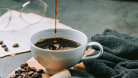 A person pours coffee from a black pitcher into a white cup. Coffee beans and papers are on the table in the background.