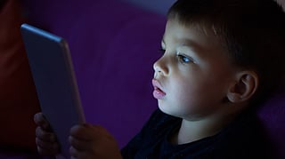 A boy sits in a dim room, focused on a glowing tablet screen.