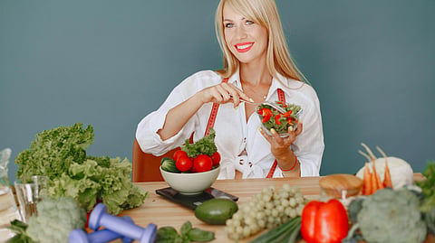 A woman sits at a table with salad, fruits, vegetables, and dumbbells, showing a healthy lifestyle.