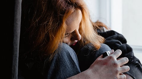 A young person sits by a window holding a cigarette and looking sad.