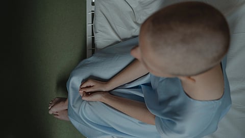 A person with a shaved head sits on the edge of a hospital bed.