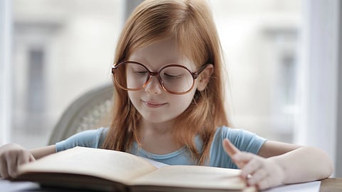 A young girl with glasses smiles while reading a book at a table.
