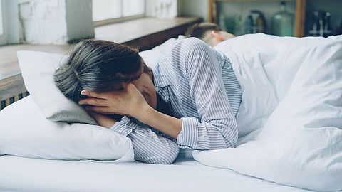 A woman lies in bed looking upset while her partner sleeps beside her.