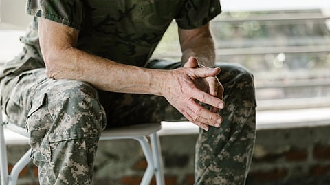 An older veteran in camouflage sits with hands clasped on a chair.