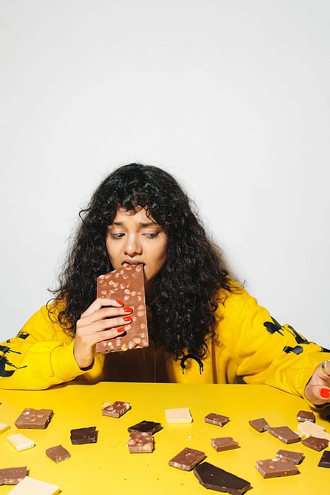 Woman biting into a milk chocolate hazelnut bar with chocolate pieces scattered on a yellow surface