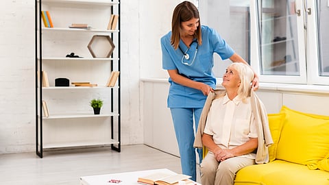 A home care nurse helps an elderly woman sit comfortably on a couch.