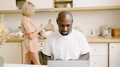 A man works on a laptop while a woman prepares food nearby.