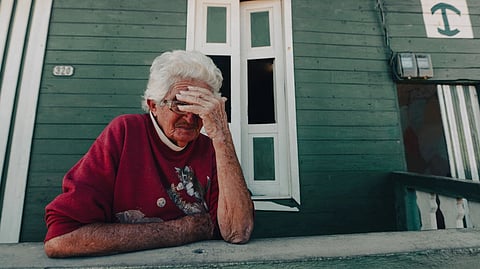 An elderly woman sits outside with her hand on her forehead.