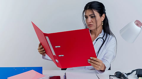 A healthcare worker looks concerned while reviewing a red file at her desk.