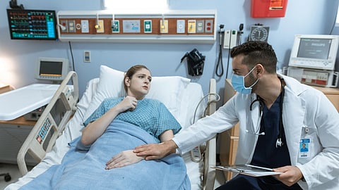 A doctor checks on a patient while reviewing a clipboard in a hospital room.
