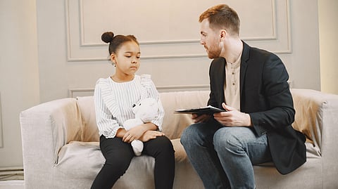 A young girl sits on a sofa holding a teddy bear.