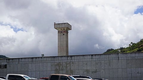 A prison watchtower stands behind a high security wall under a cloudy sky.