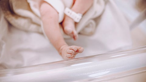 A newborn baby's tiny feet rest in a clear hospital bassinet with an ID band on one ankle.