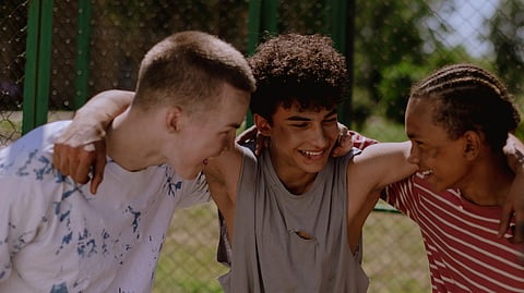Three teenage boys smile together outdoors with their arms around each other.