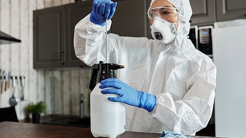 A person in protective gear sprays a kitchen surface with a cleaning bottle.