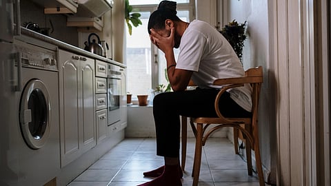 A person sits alone in a kitchen with their head in their hands, looking distressed.