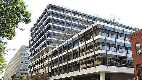 A modern glass office building with white bands, a rooftop terrace, and nearby brick and office buildings under a clear sky.