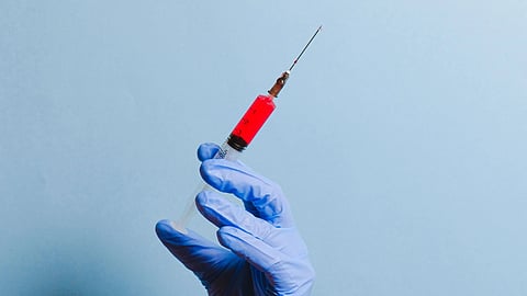 A gloved hand holds a syringe filled with red liquid against a blue background.