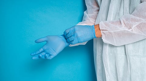 A healthcare worker puts on a blue disposable glove while wearing a protective gown.