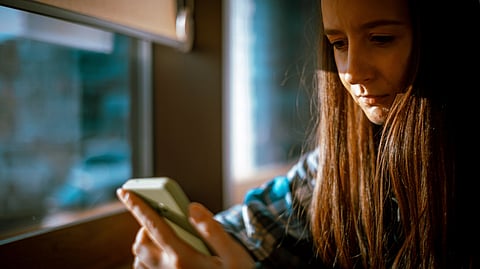 A young woman with long brown hair sits by a window.