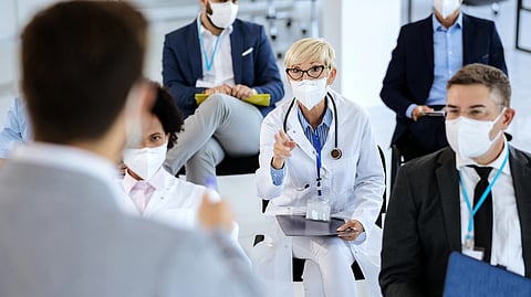 A masked doctor speaks during a medical conference as healthcare professionals listen.