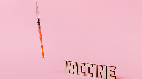 An insulin syringe stands beside the word "VACCINE" on a pink background.