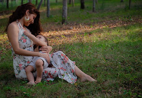 / Pexels	Mother sitting outdoors on grass, breastfeeding her toddler in a wooded park setting
