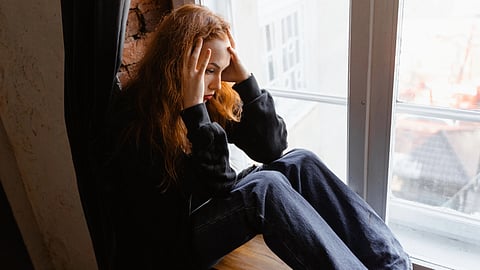 A person sitting by a window with their head in their hands, looking distressed.