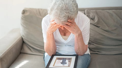 An elderly person sits on a sofa holding a framed photo, with their head in their hands, expressing grief or sadness.