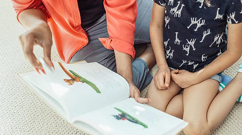 An adult reads a picture book with a child sitting nearby.