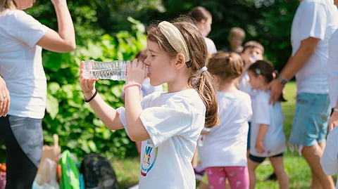 Girl drinks water from a bottle outdoors in a sunny park.