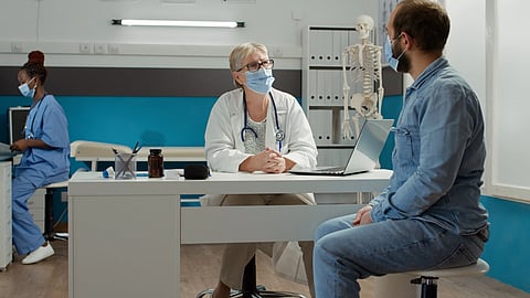 Masked female doctor consults with a seated male patient, with a nurse and skeleton model nearby.