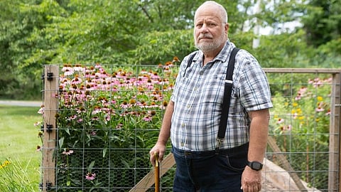 An older man with a gray beard stands with a cane in a garden.