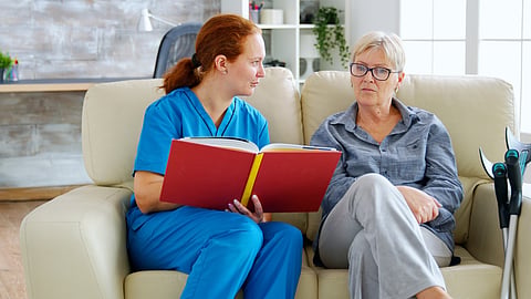 A healthcare worker in blue scrubs talks with an elderly woman on a sofa, with crutches beside her.