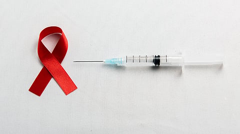A red awareness ribbon sits next to a medical syringe with a needle against a plain white background.