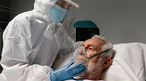A PPE-clad healthcare worker adjusts an oxygen mask for an elderly patient in a hospital bed.