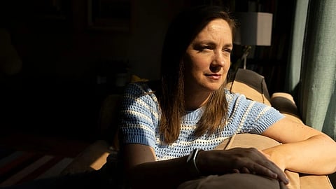 A woman sits thoughtfully on a couch, illuminated by sunlight from a window.
